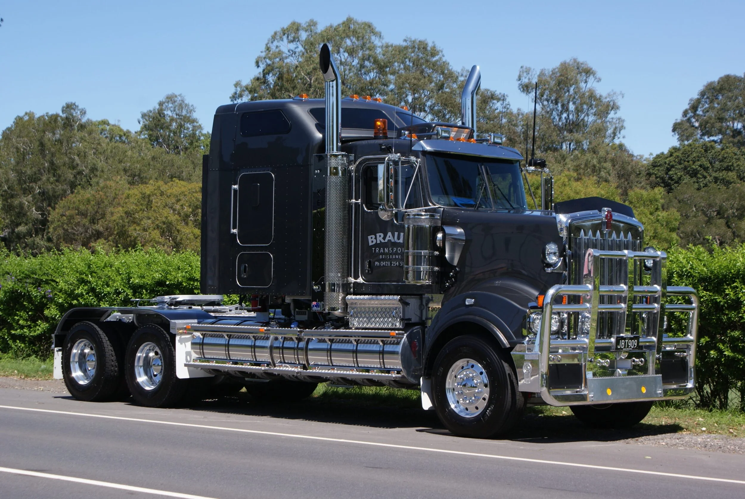 Black semi-truck parked on the side of a road with greenery and trees in the background.