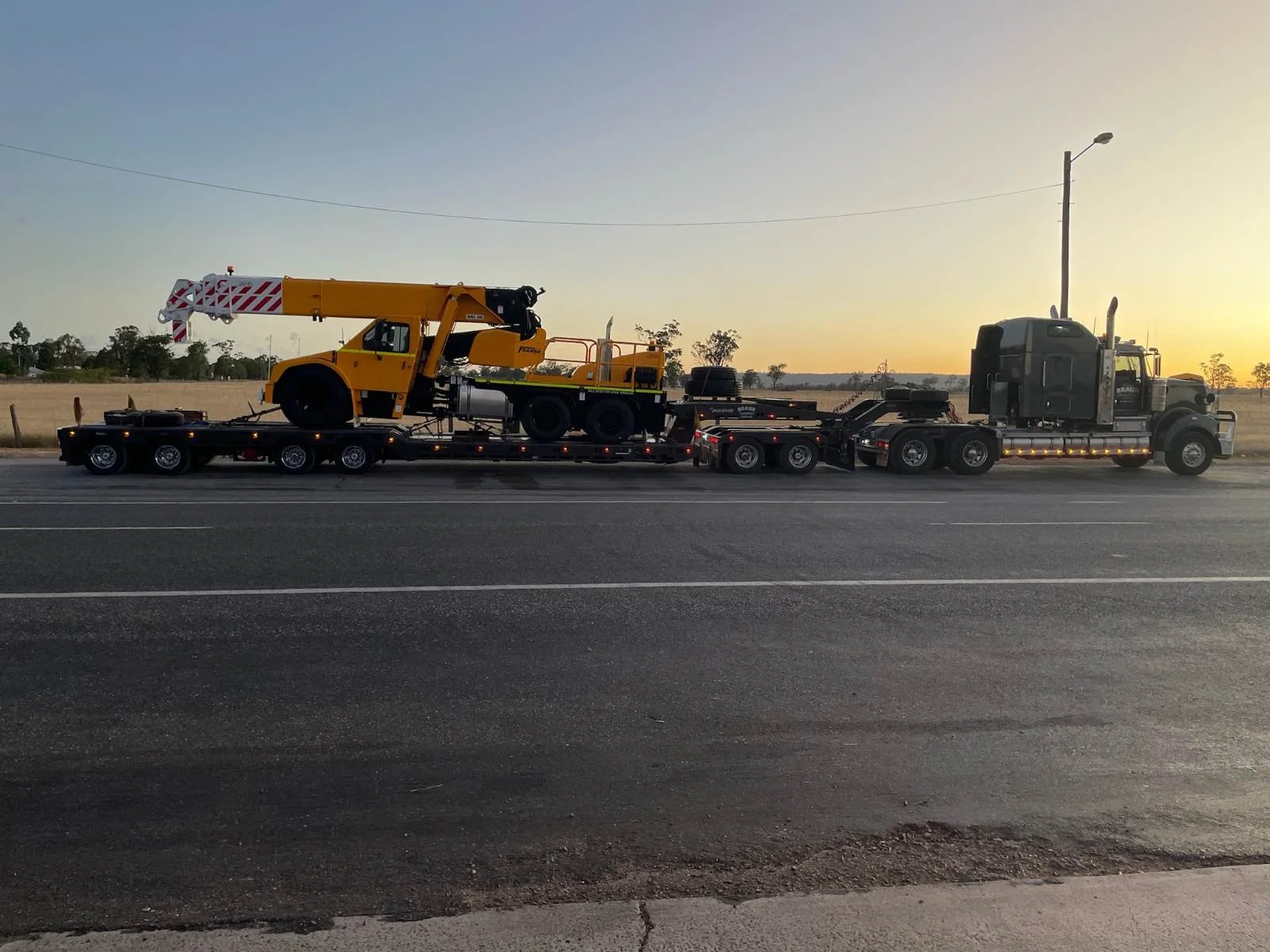 A large semi-truck with a flatbed trailer carrying a yellow crane is parked on the side of a road during sunset. The background features open fields and a few scattered trees.