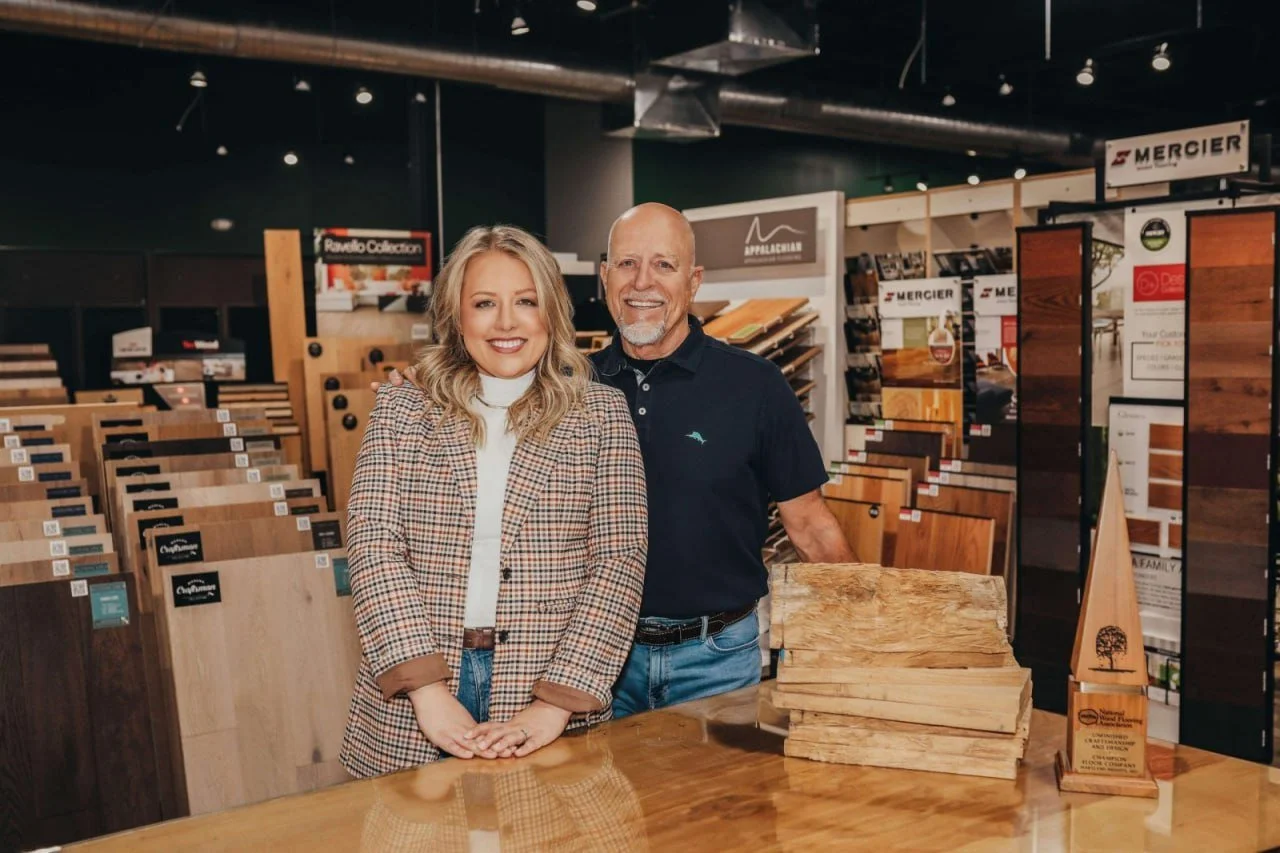 A smiling woman and man standing inside a flooring store, surrounded by wood flooring samples, with a stack of wood planks on the table in front of them.