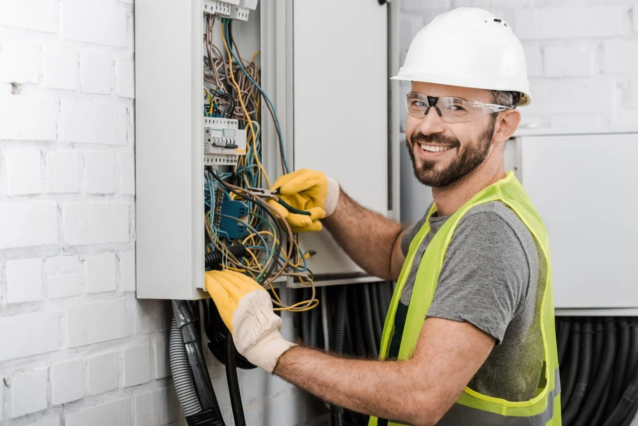 A smiling electrician with a white hard hat, safety glasses, and yellow gloves working on an electrical panel.
