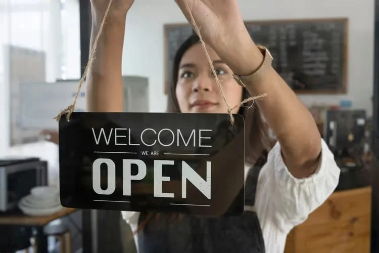 Woman hanging a sign that says 'Welcome we are open' in a cafe or shop.