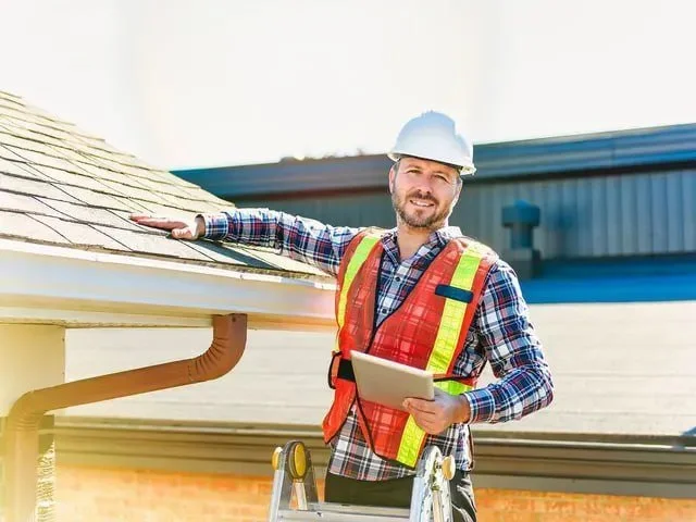 A male construction worker with a beard, wearing a white safety helmet and a red safety vest, holding a tablet, standing next to a house with a sloped roof during daytime.