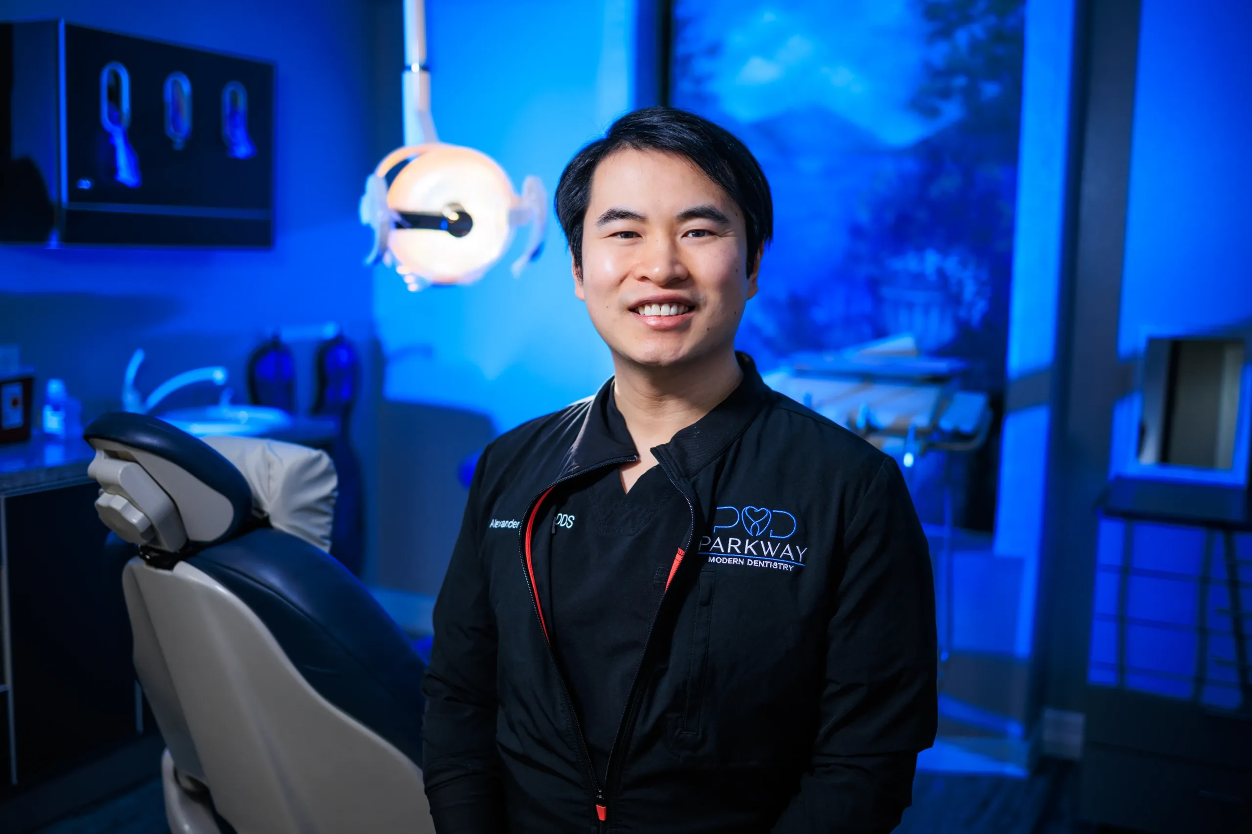 A smiling man dressed in black dental scrubs standing inside a modern dental office with blue lighting, dental chair, and dental equipment in the background.