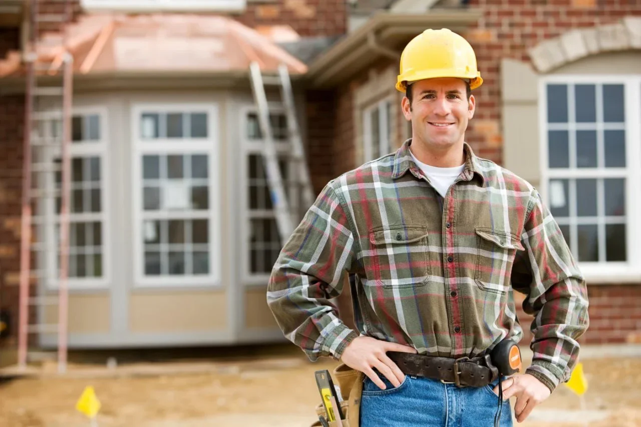 A construction worker in a plaid shirt and yellow hard hat standing in front of a house under construction.