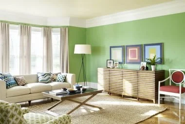 Living room with green walls, three framed pictures, a beige sofa with decorative pillows, a coffee table, a white floor lamp, a sideboard with picture frames and a plant, and a red and white chair.