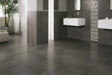 Modern bathroom with gray tiled floor, dark wall tiles, two white wall-mounted sinks, and a potted plant in the corner.