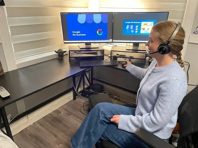 A woman with a headset working at a dual-monitor computer setup with Google My Business screens, sitting in a home office.