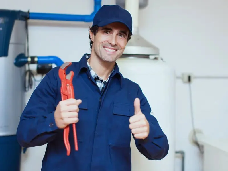 A smiling male plumber in a blue uniform and cap, holding a red pipe wrench in one hand and giving a thumbs up with the other, standing in a plumbing or utility room.