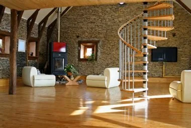 Modern living room with stone walls, wooden ceiling beams, a spiral staircase, beige couches, and electronic devices.
