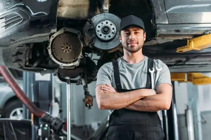 A mechanic standing with arms crossed under a lifted car in a garage, holding a wrench, with the car's brake disc and suspension visible.