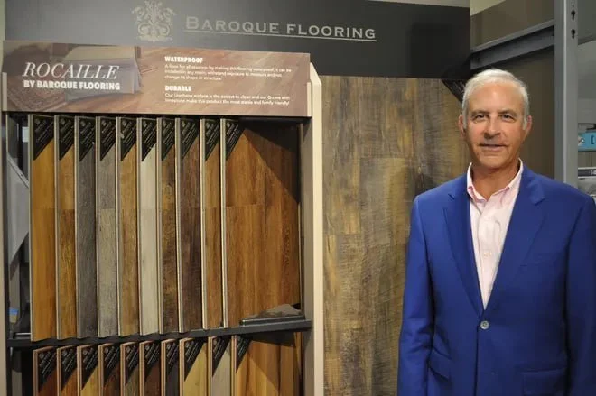 A man in a blue blazer and white shirt standing next to a display of wood flooring samples at a flooring store.