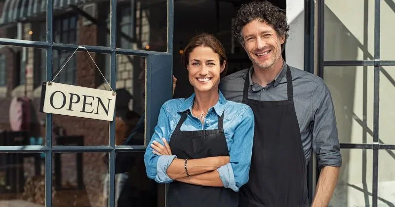 Smiling couple with aprons standing in front of a cafe with an 'Open' sign in window.