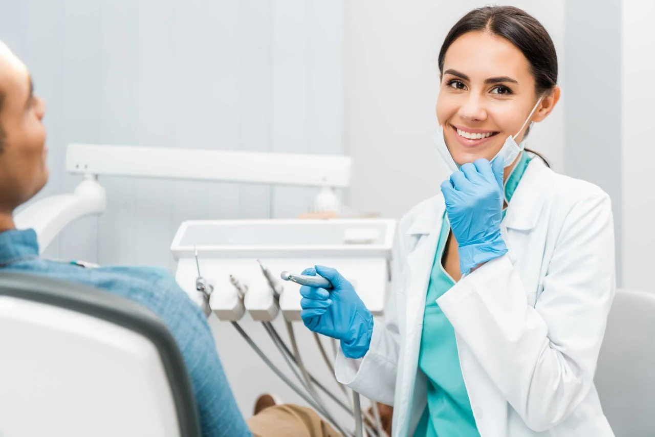 Female dentist smiling at the camera in a dental office, wearing a white coat, blue gloves, and a face mask pulled down, holding dental tools near a patient in an exam chair.