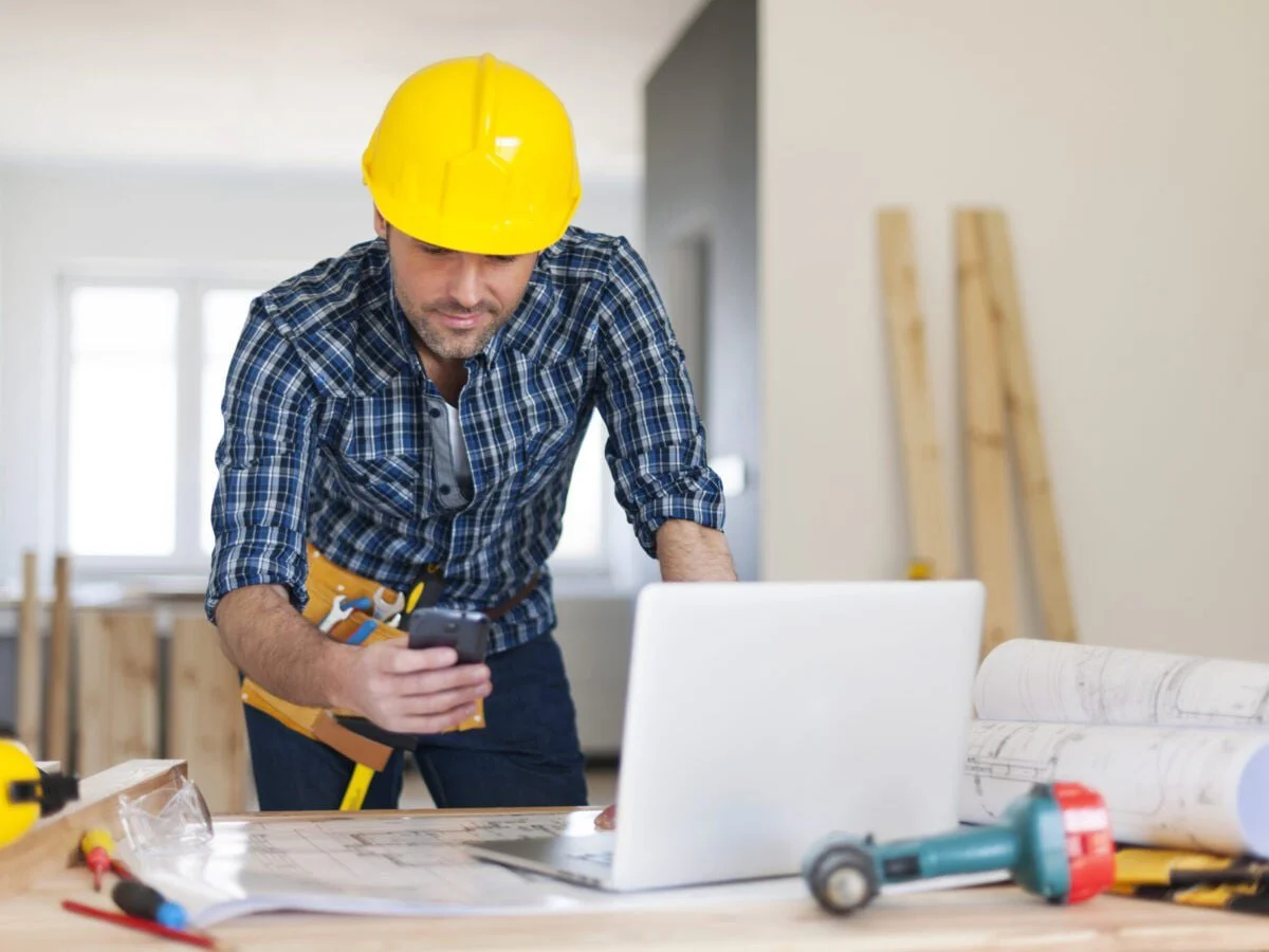 A man wearing a yellow construction helmet and plaid shirt looking at his phone while working at a desk with tools, a laptop, and blueprints in a construction site or renovation setting.