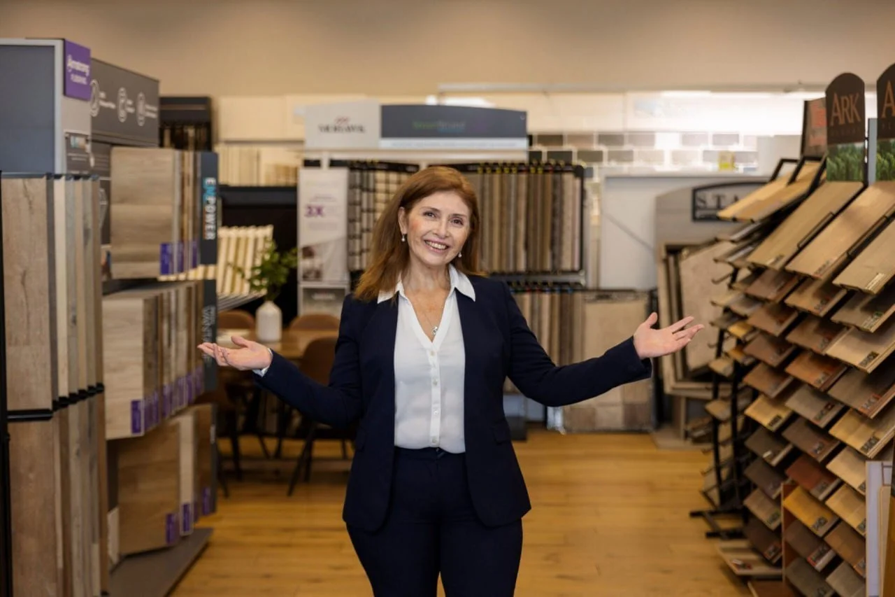 A woman in business attire standing in a flooring store, smiling and gesturing with open arms, with display shelves of wood and tile flooring samples behind her.