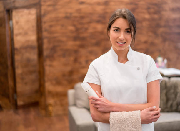 A young woman wearing a white chef's coat and holding a beige towel, standing in a kitchen with a wooden wall background.