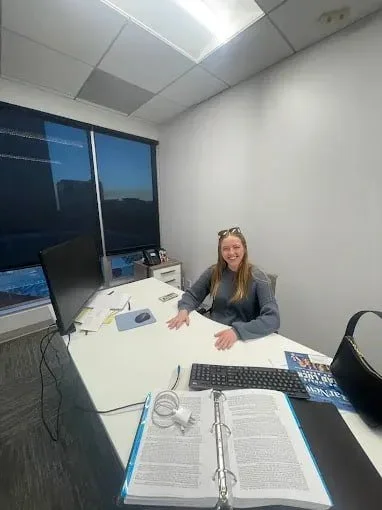 Woman sitting at a desk in an office, smiling at the camera, with a computer monitor, keyboard, open binder, and a black bag on the desk.