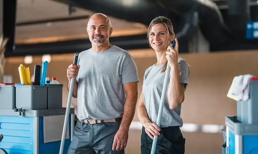 Two cleaning staff members, a man and a woman, posing with mops in a well-lit, modern building.