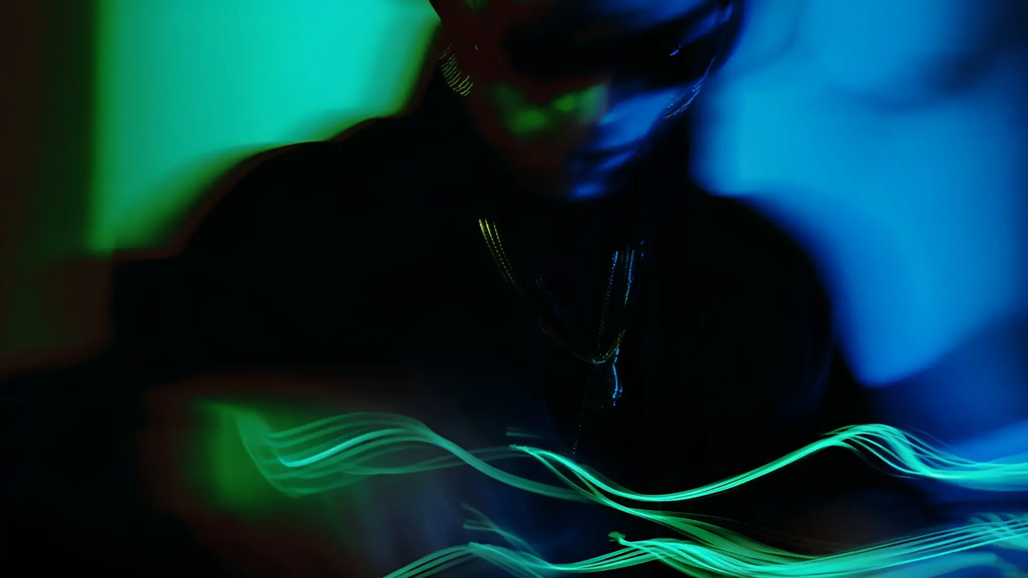 Dark image with abstract colored light streaks in green and blue, and a blurry silhouette of a person wearing sunglasses and a chain, creating a mysterious and vibrant visual effect.