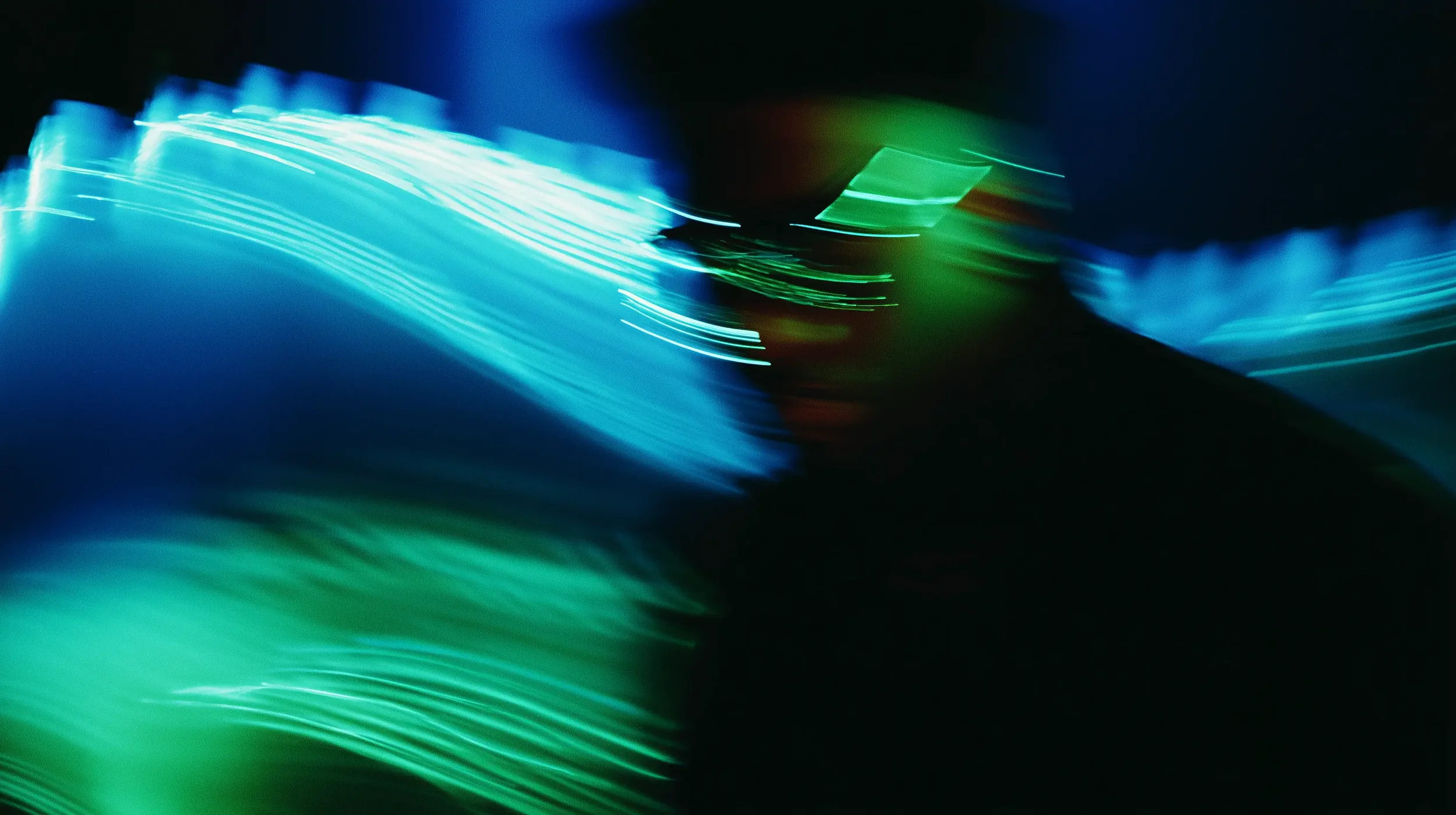 Abstract image with streaks of blue, green, and hints of red and black, resembling light trails or long exposure light photography.