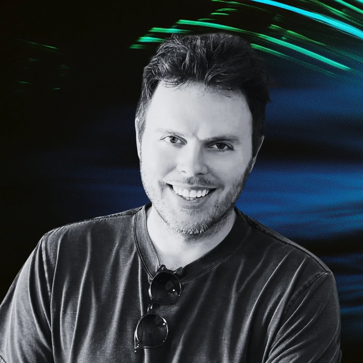 A smiling man with dark hair and a beard, wearing a dark T-shirt and sunglasses hanging from his shirt, in front of a vibrant backdrop with green and blue streaks of light.