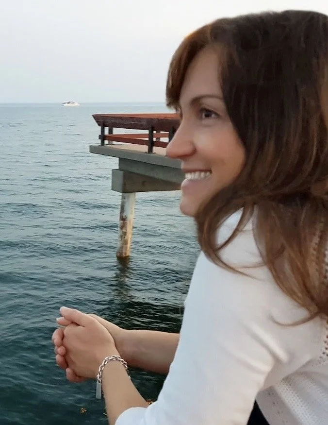 A woman with brown hair smiling, sitting by the water near a pier, with a boat in the distance.