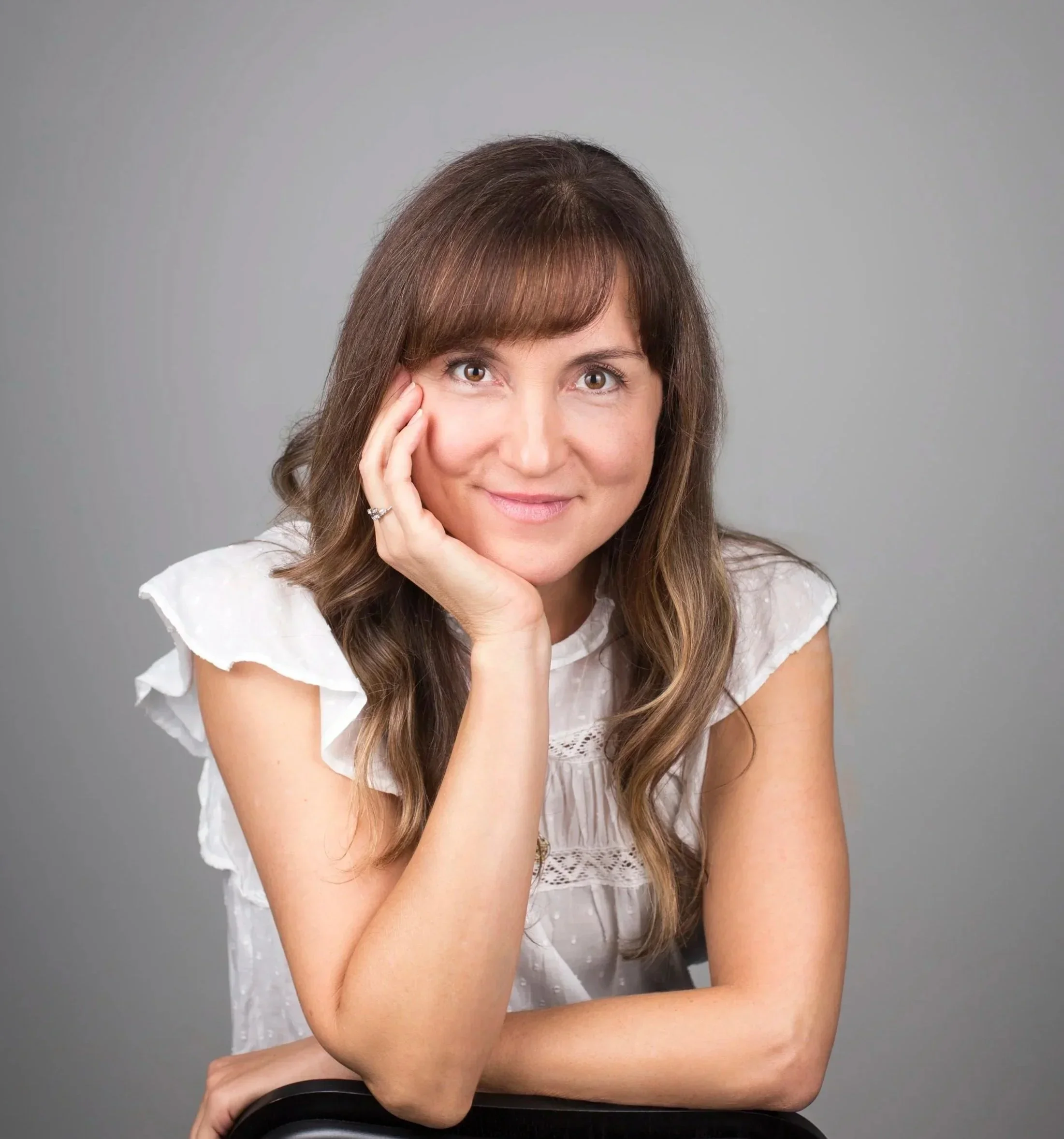 A woman with brown hair and a white blouse resting her chin on her hand, smiling at the camera against a plain gray background.
