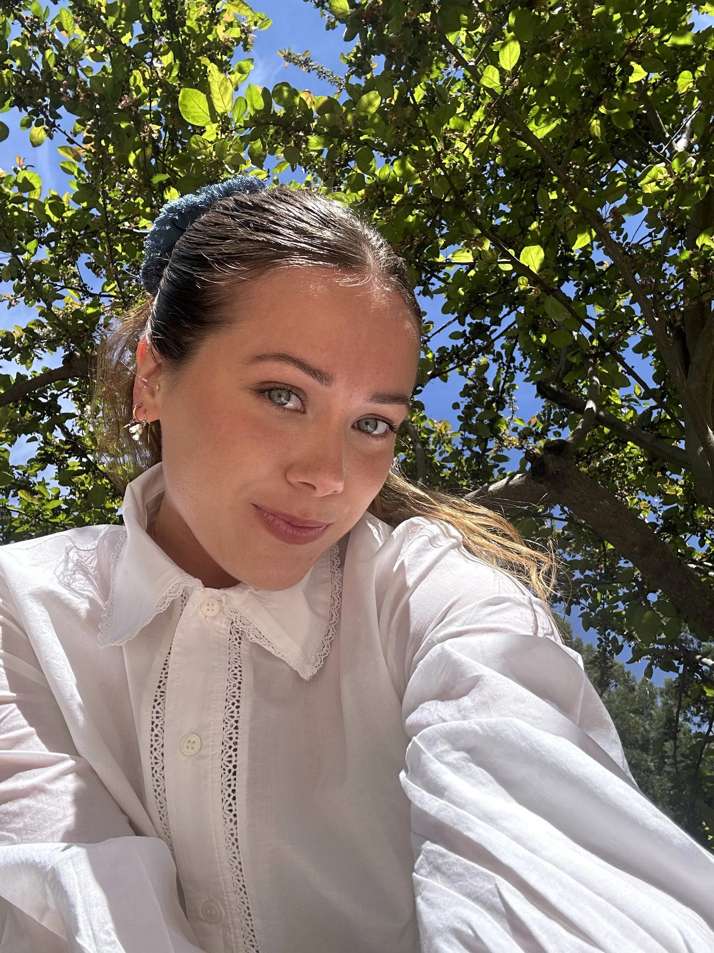 A woman with light skin, brown hair, and blue eyes taking a selfie outdoors, with a background of leafy green trees and a clear blue sky.