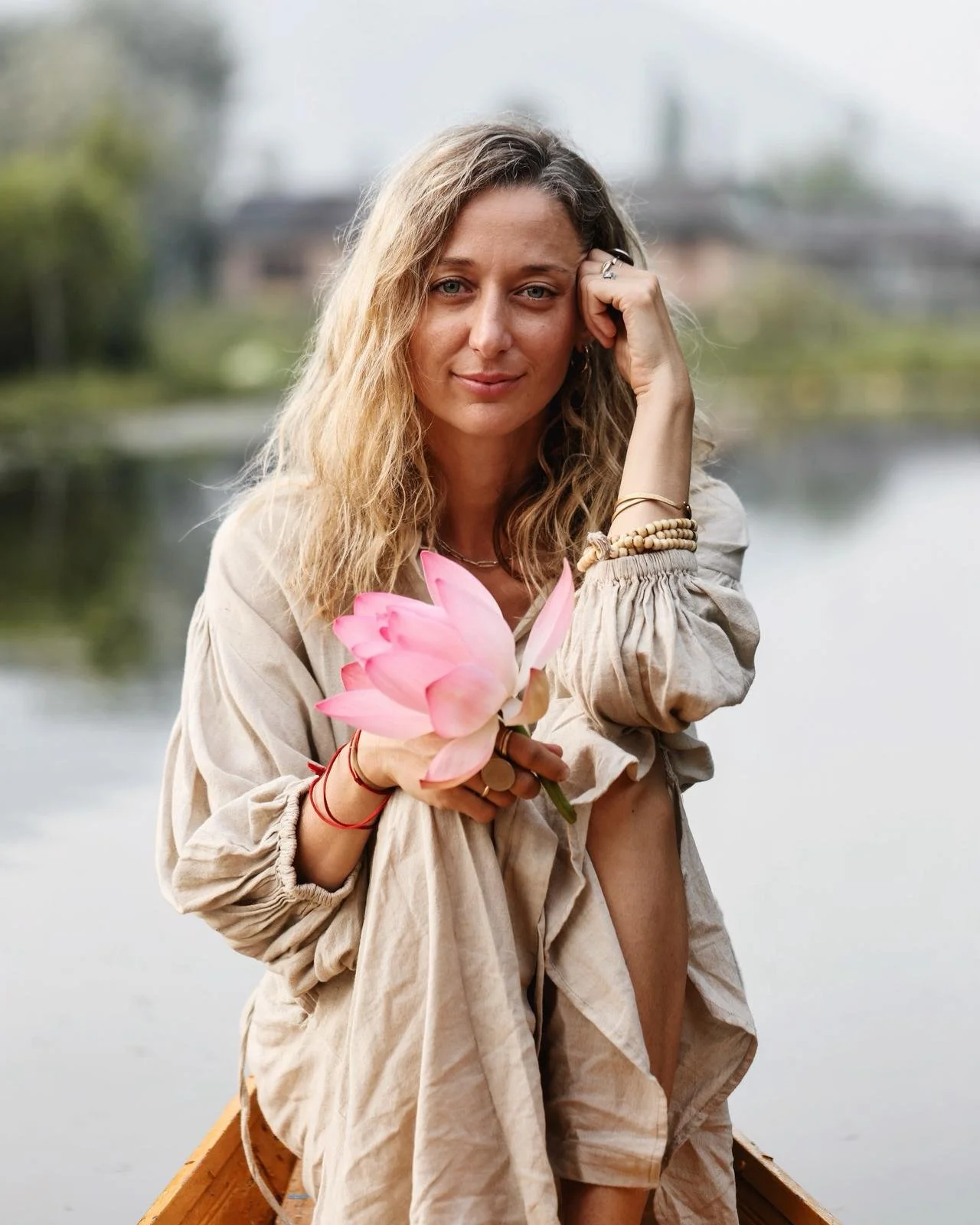 Nina Karnikowski is one of our teachers at the immersion. A woman with curly blonde hair holding a pink lotus flower while sitting in a boat on a river, with blurred greenery and buildings in the background.