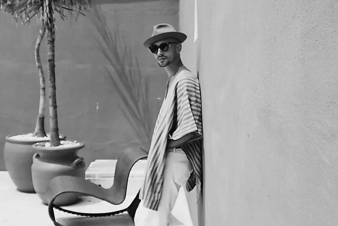 Timo Granzotti, lead teacher is wearing a beige wide-brim hat, sunglasses, a striped shirt, and light-coloured pants stands outdoors against a peach-colored wall near potted plants.