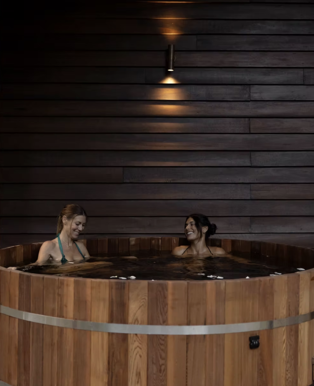 Two women relaxing in a round wooden hot tub indoors, smiling and enjoying each other's company.