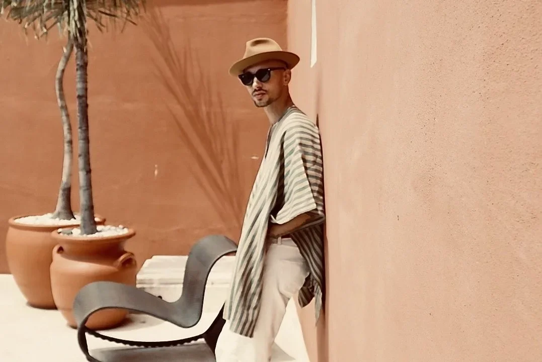 Timo Granzotti, lead teacher is wearing a beige wide-brim hat, sunglasses, a striped shirt, and light-coloured pants stands outdoors against a peach-colored wall near potted plants.