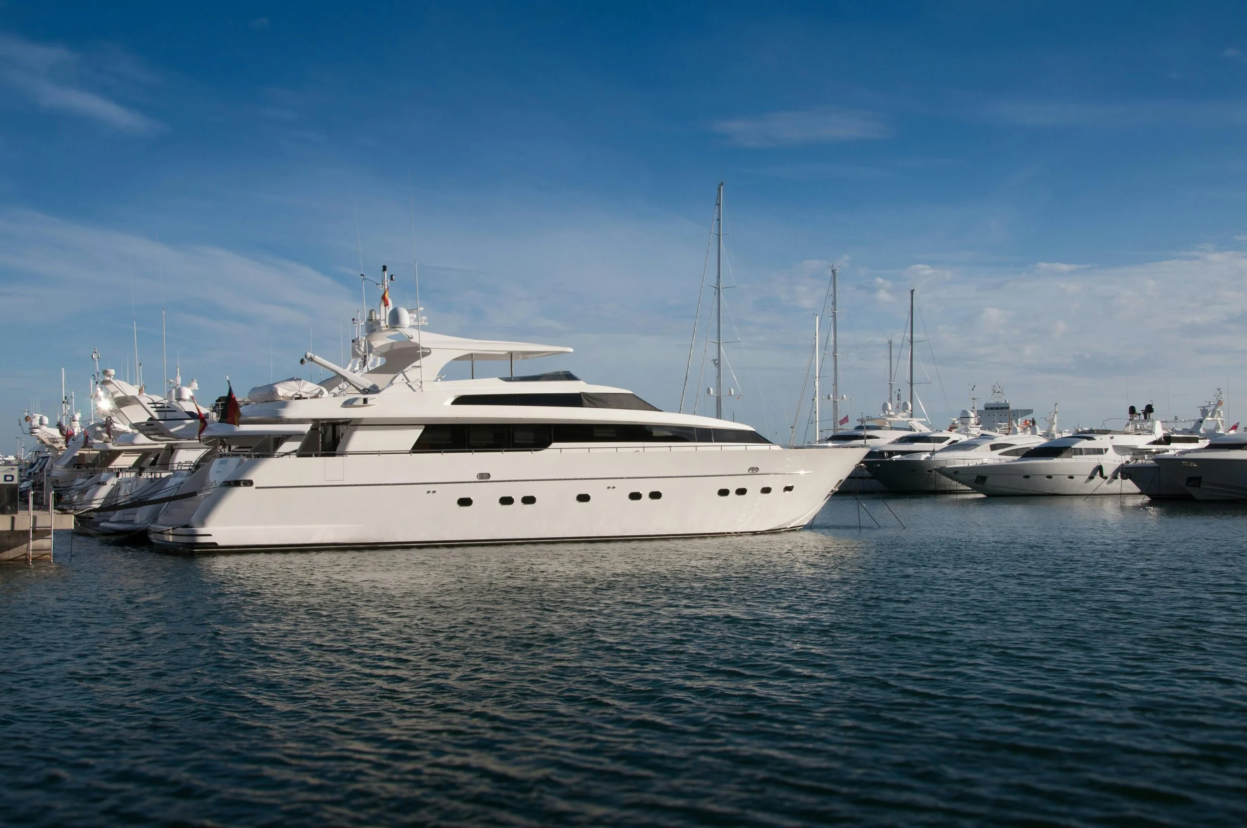 A row of luxury white yachts docked at a marina on a clear day with blue sky and calm water.