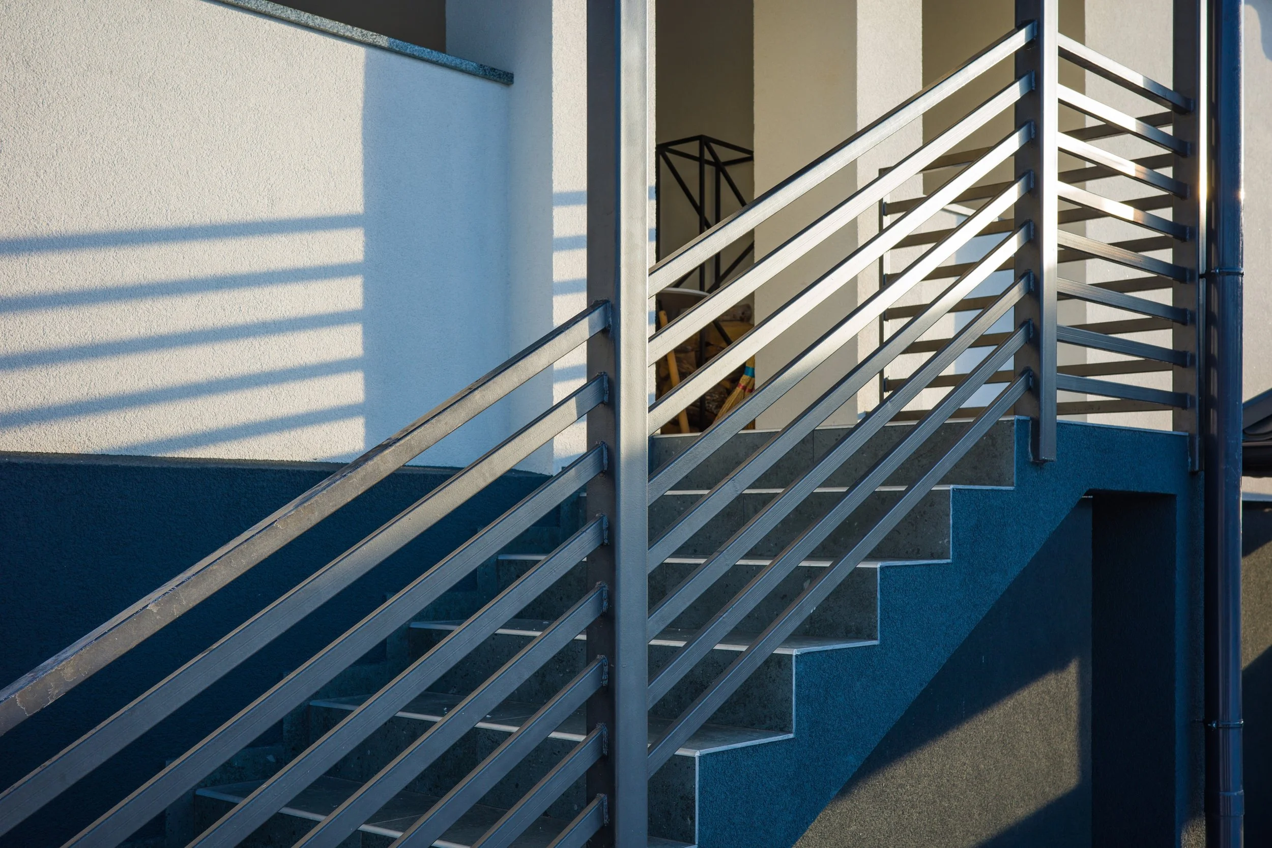 Exterior metal staircase with railing leading up to a doorway, shadows cast on white wall, modern building design.
