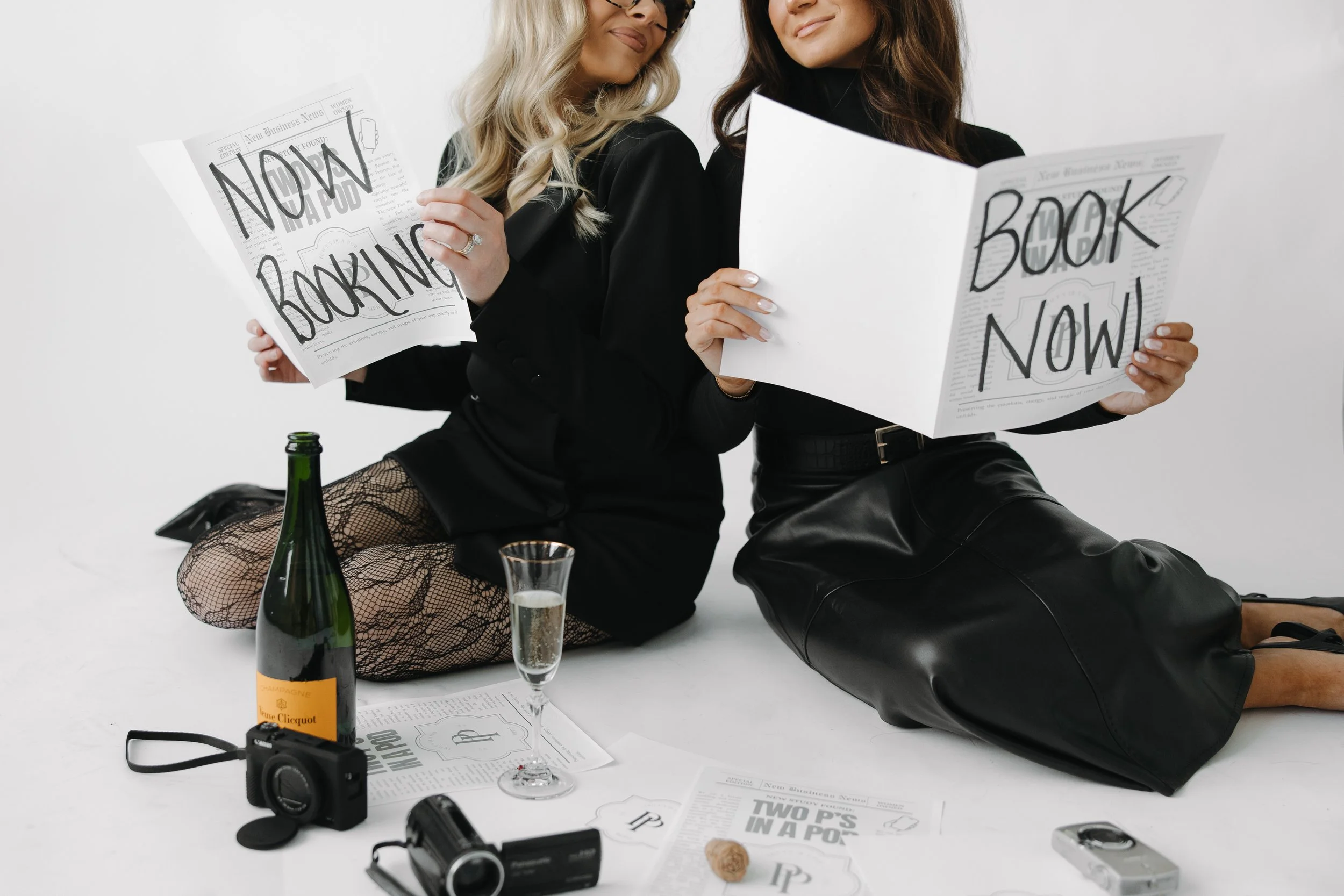 Two women sitting on the floor holding newspapers that say "BOOK NOW". One has blonde hair, the other has brunette hair. Items on the floor include a champagne bottle, a glass of champagne, a camera, a magazine, and a phone.