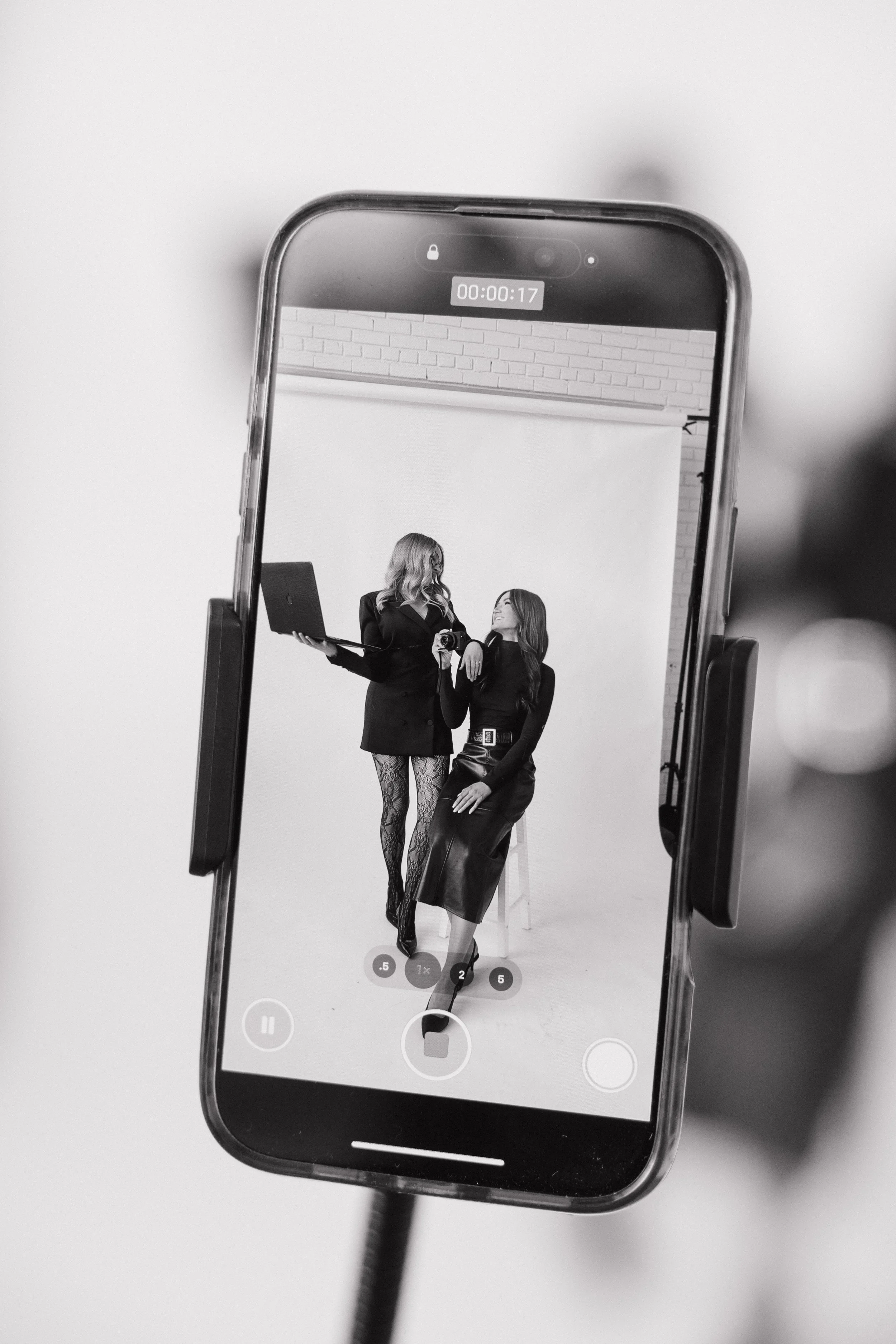 A phone is capturing a black and white photo of two women in a studio, one standing with a laptop and the other sitting on a stool, both smiling and dressed in fashionable black outfits.