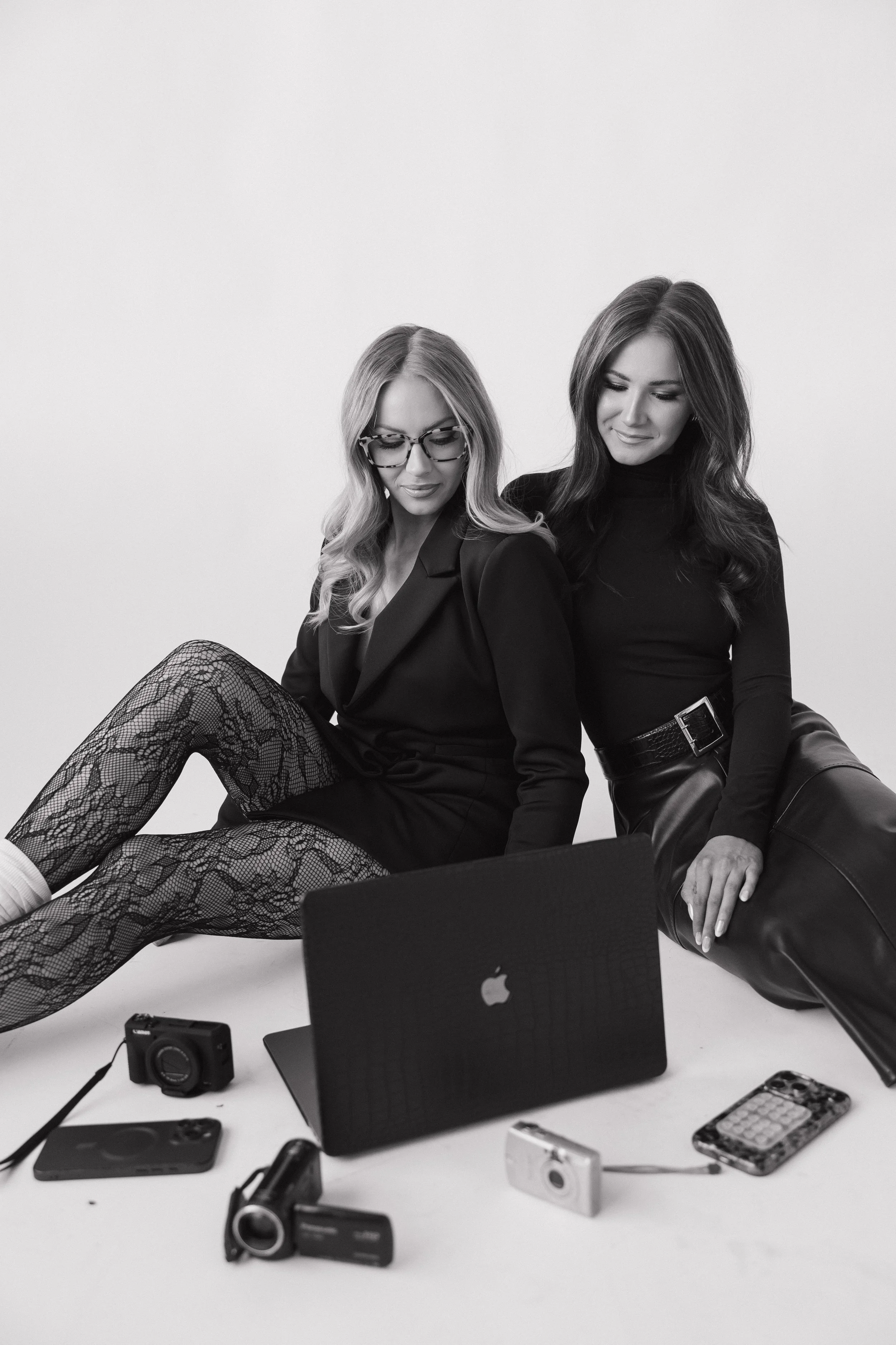 Two women sitting on the floor with a laptop and various cameras and a phone, in a studio with a white background.