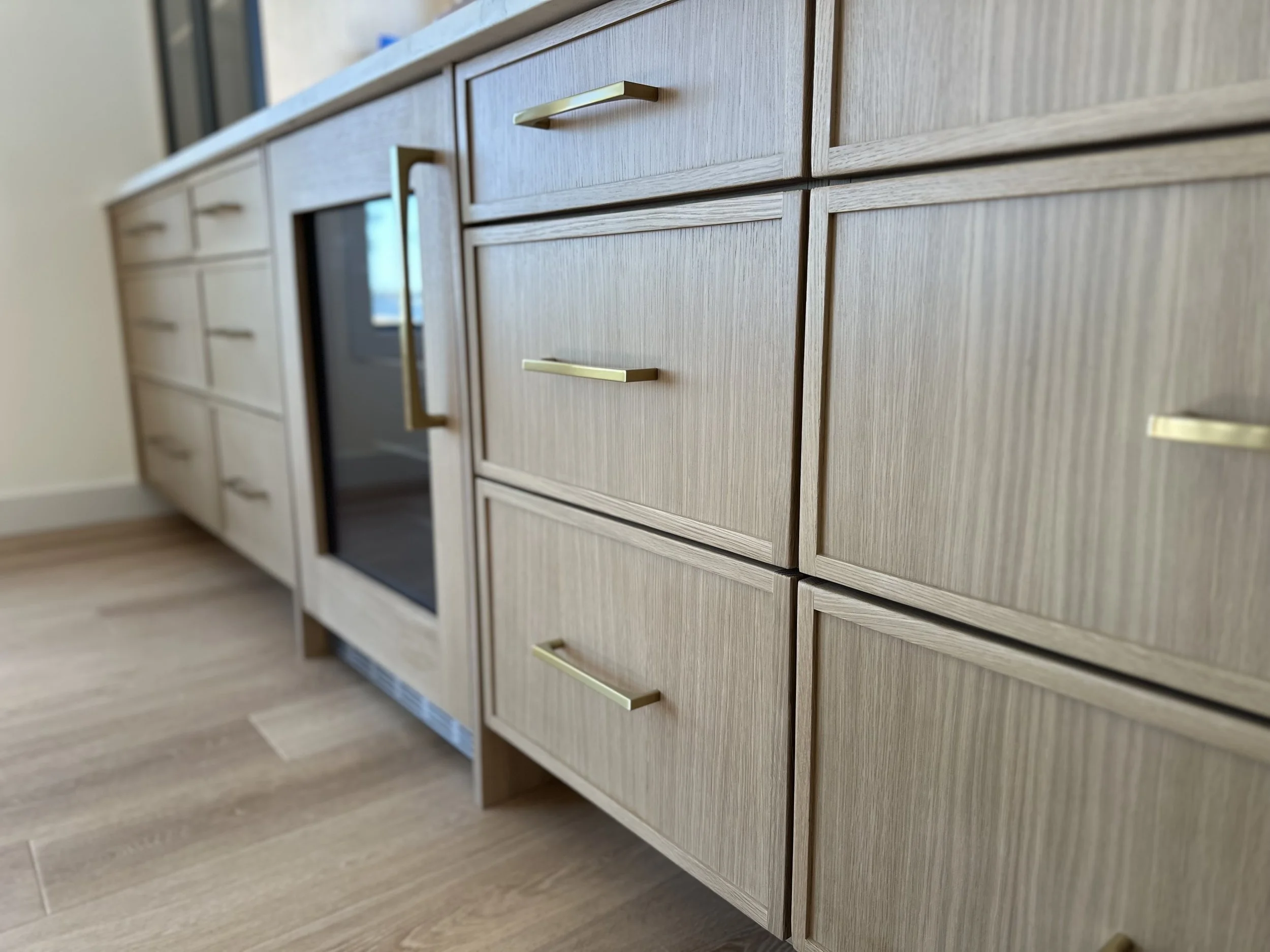 Close-up of light wood kitchen cabinets with gold handles, including drawers and a built-in oven, in a modern kitchen with hardwood flooring.