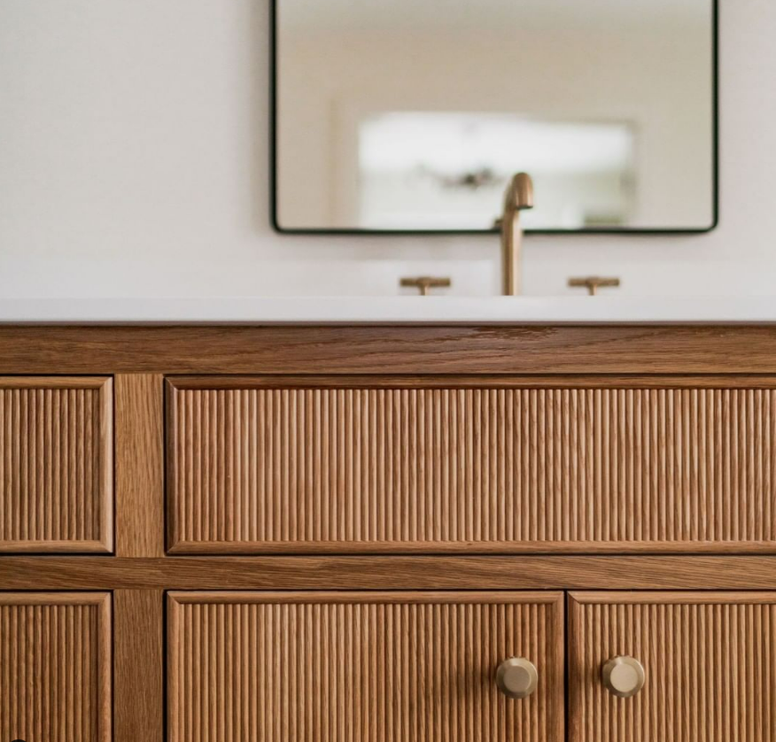 Wooden bathroom vanity with ribbed cabinet doors, a mirror, and a gold faucet.