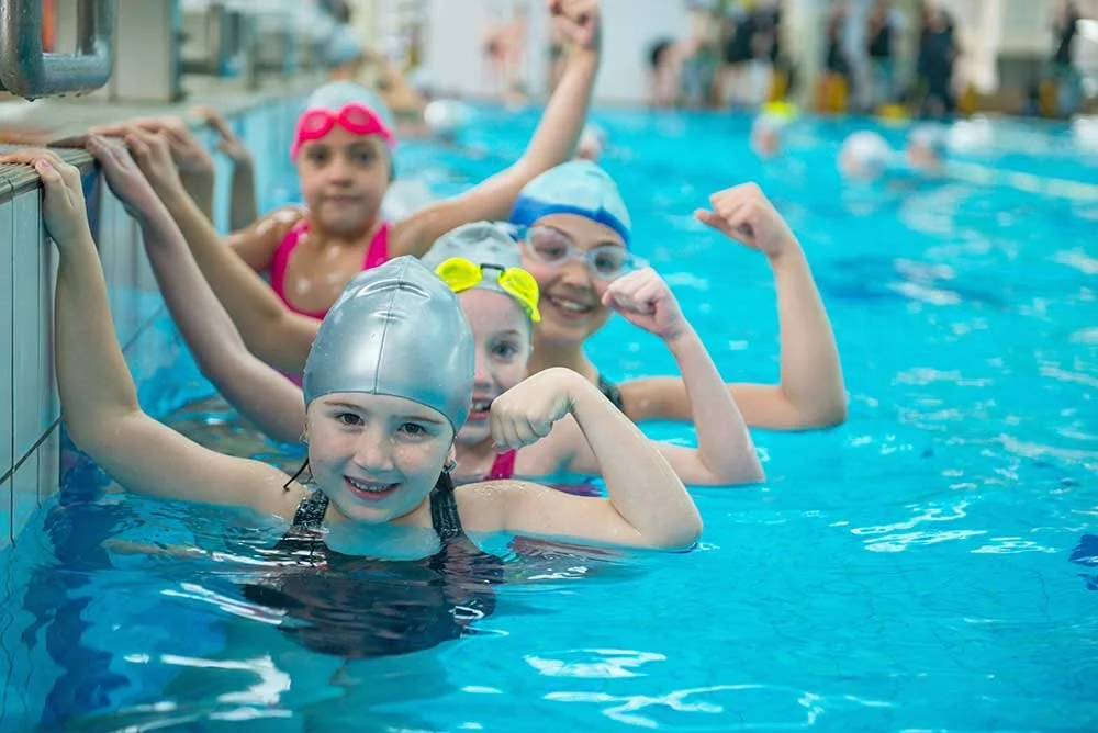 Group of children in a swimming pool wearing swim caps and goggles, smiling and flexing arms, indicating strength and fun.