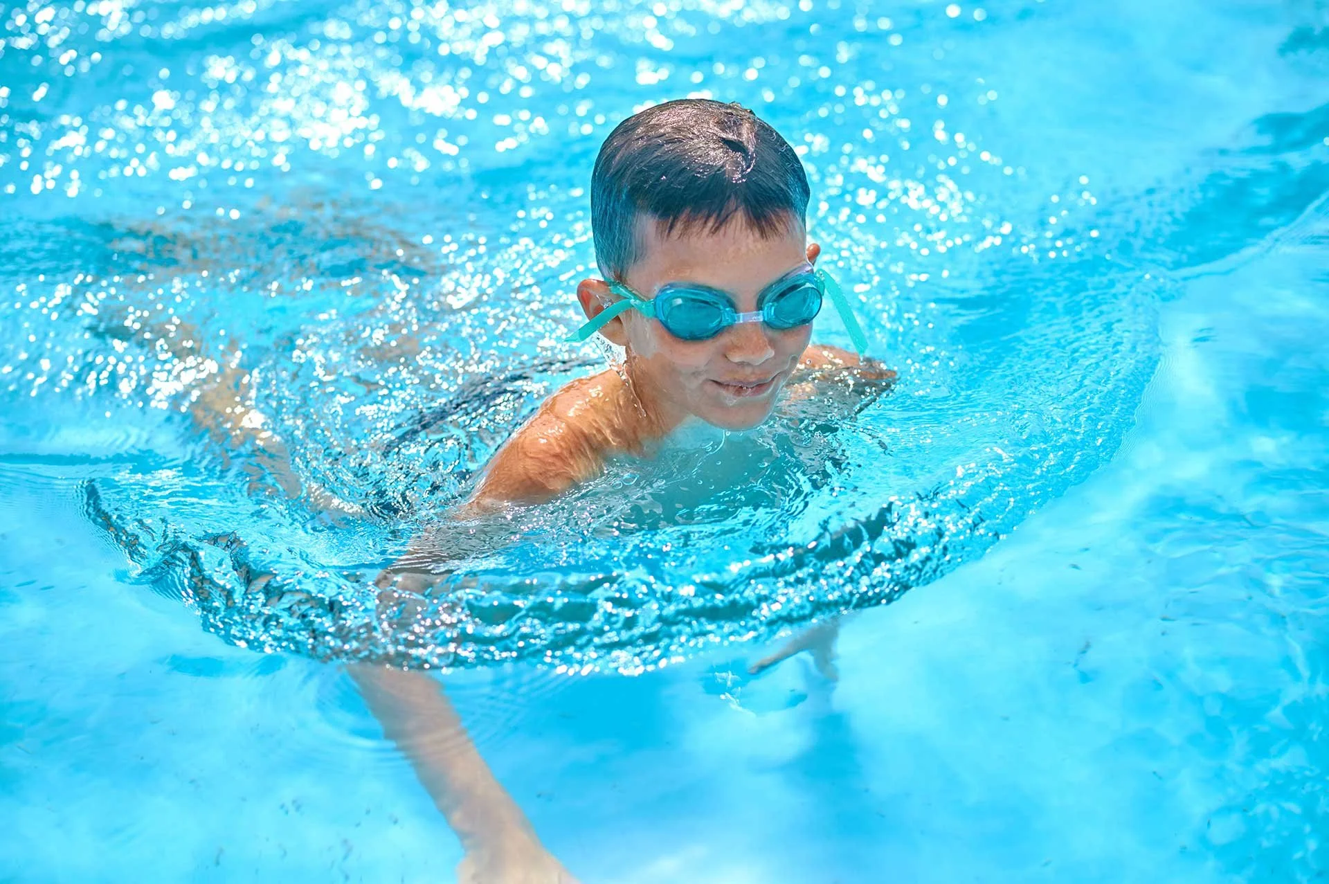 A young boy swimming in a pool wearing blue goggles.