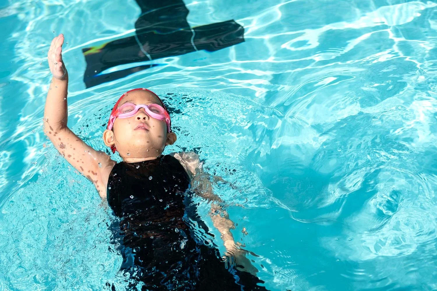 Young girl swimming in a pool, wearing pink goggles and a black swimsuit, with her eyes closed and one arm raised above her head.