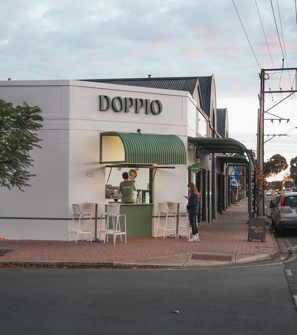 A white building with a green awning and sign that reads DOPPIO, with a woman standing on the sidewalk looking at her phone and a person working at the window counter, outside a coffee shop or café.