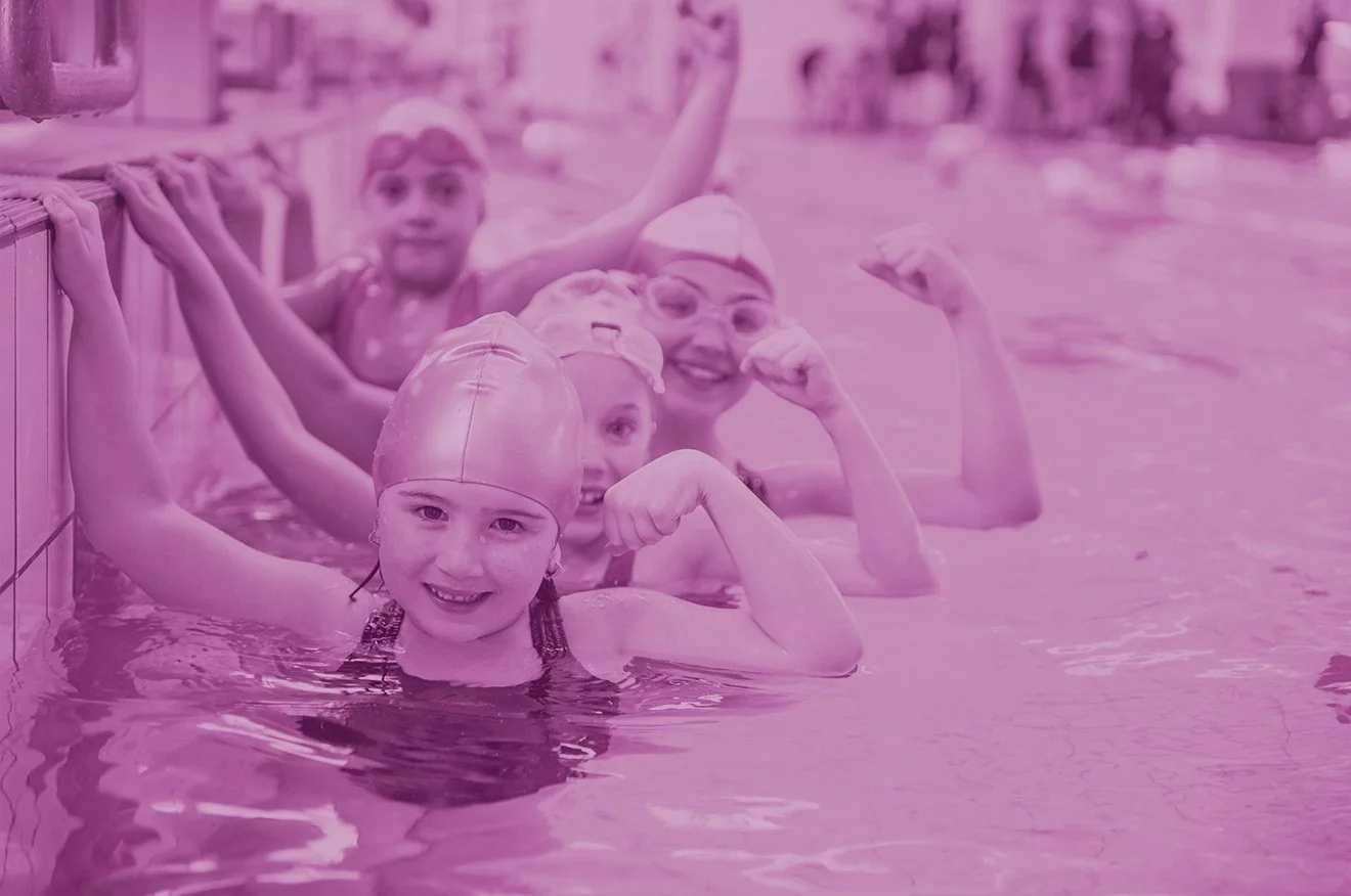 Four children and a woman in a swimming pool, smiling and flexing their muscles, wearing swim caps.
