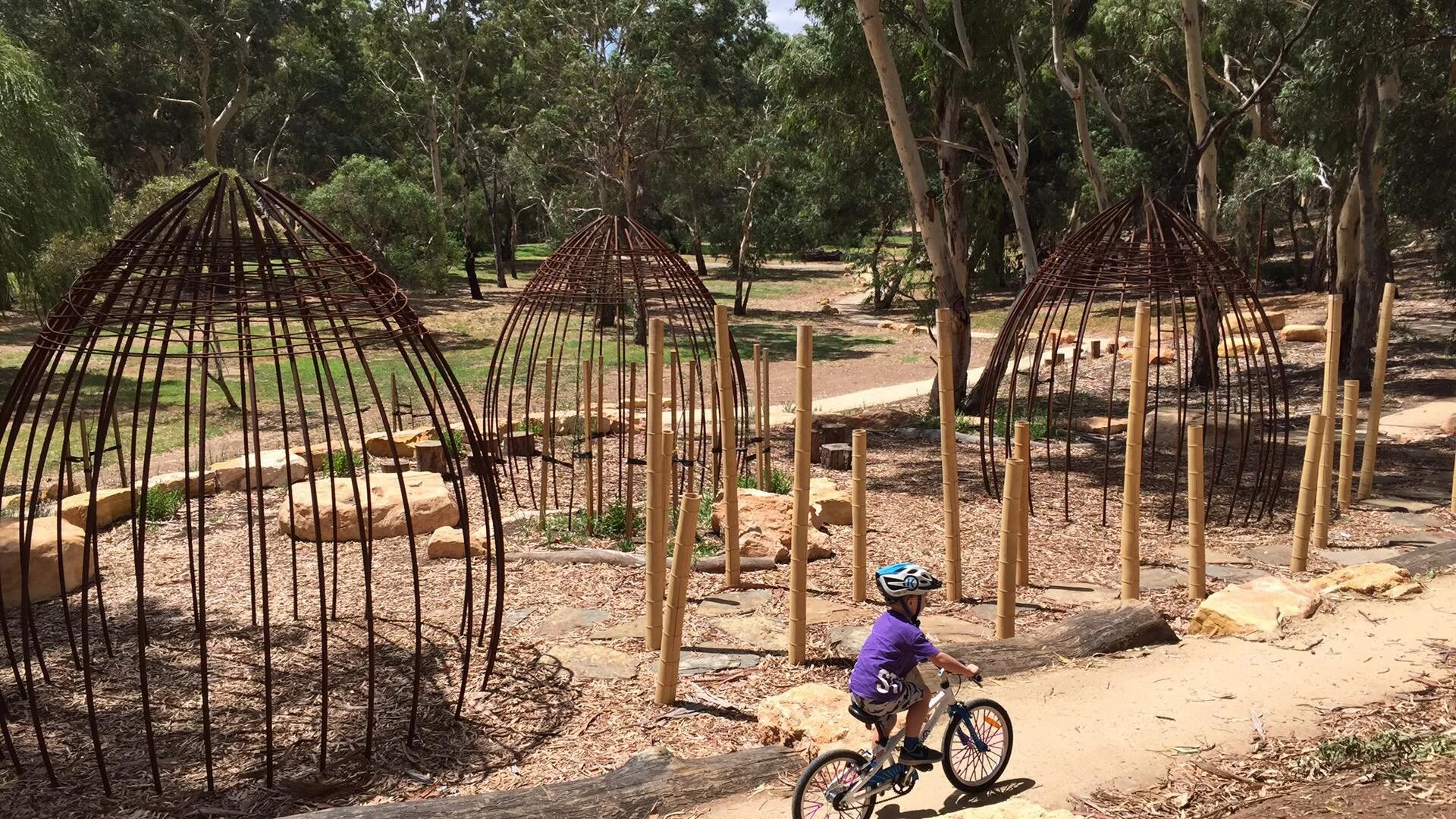 A young boy in a purple shirt and helmet rides a bicycle past a series of small, dome-shaped, metal and bamboo structures in a park with trees and dirt ground.