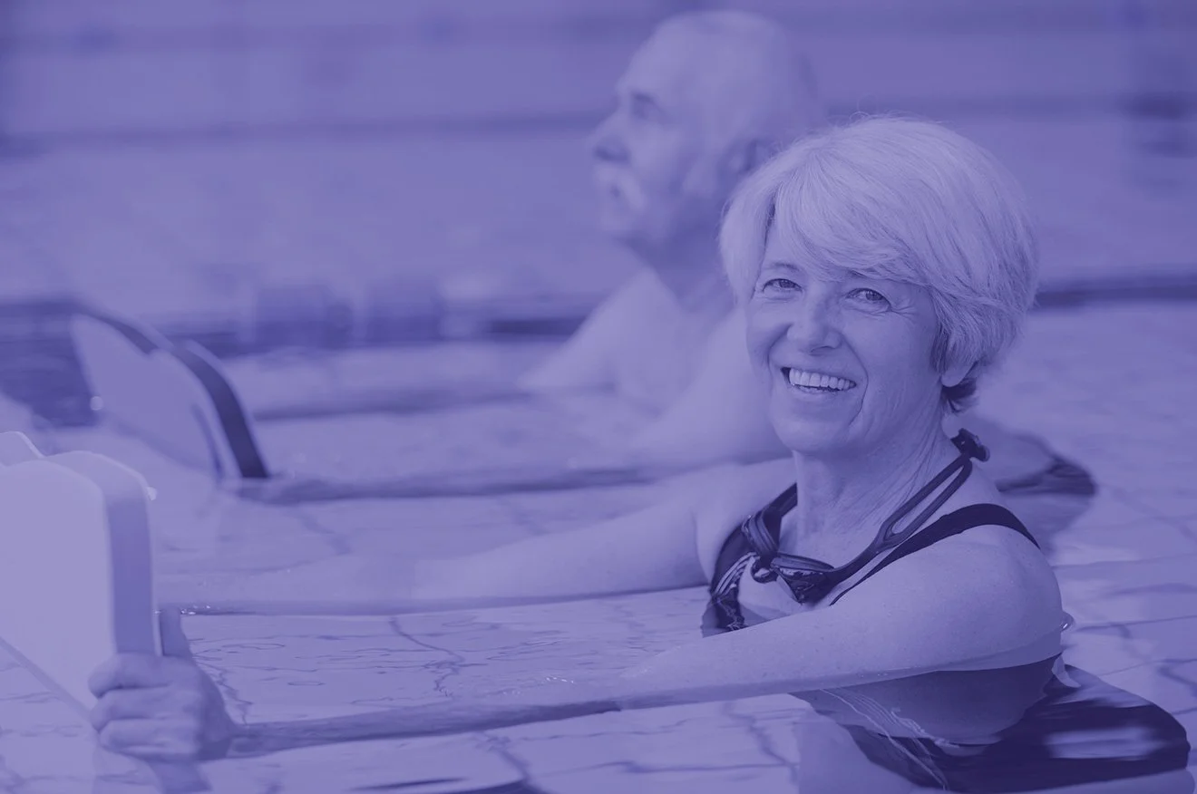 Smiling elderly woman and a man swimming in a pool, holding onto swimming aids.