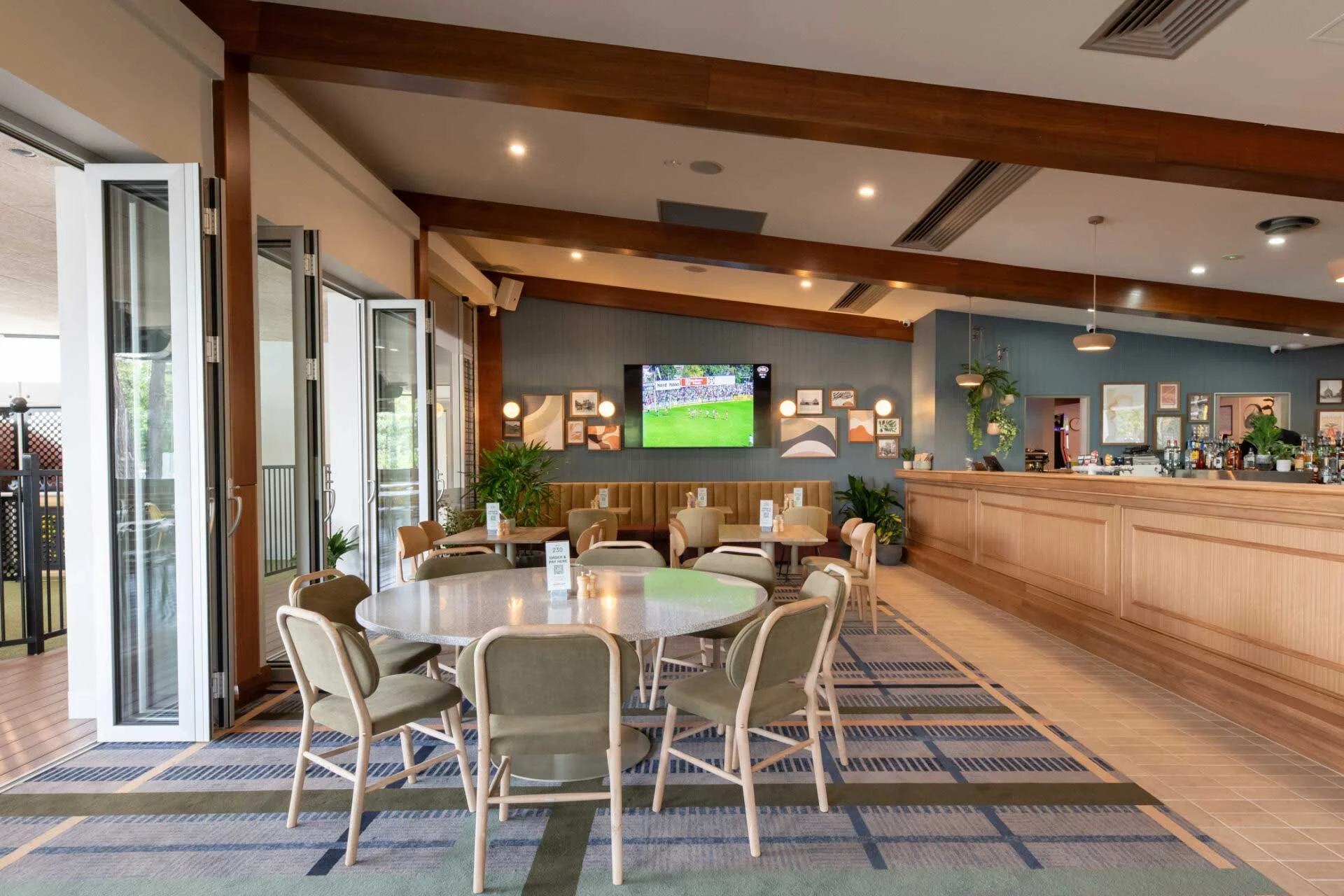 Interior of a modern restaurant or bar with round tables, beige chairs, a wooden bar counter, framed artwork on the walls, and a television showing a sports game.
