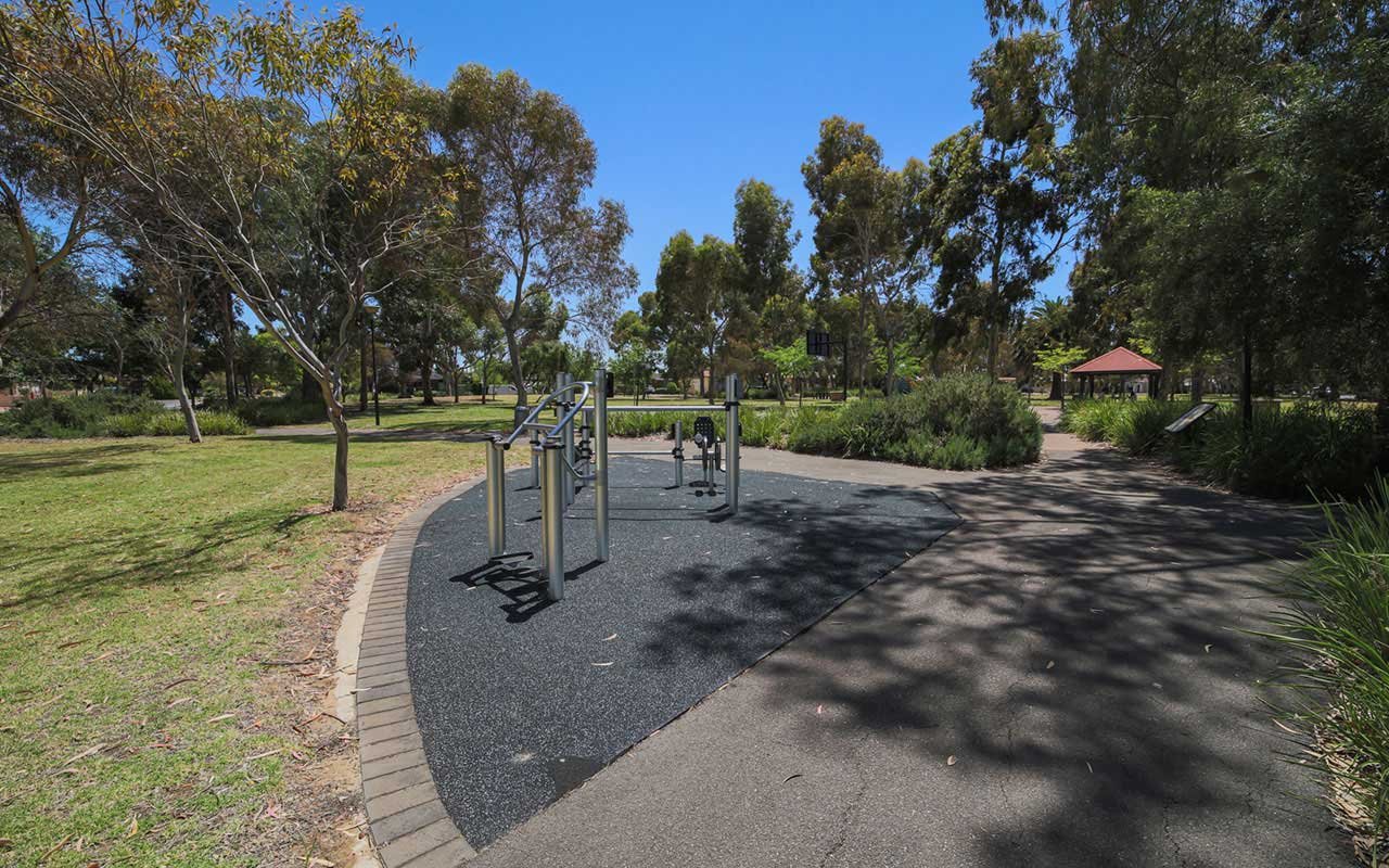 An outdoor fitness area with exercise equipment under a clear blue sky, surrounded by trees and walking paths in a park.