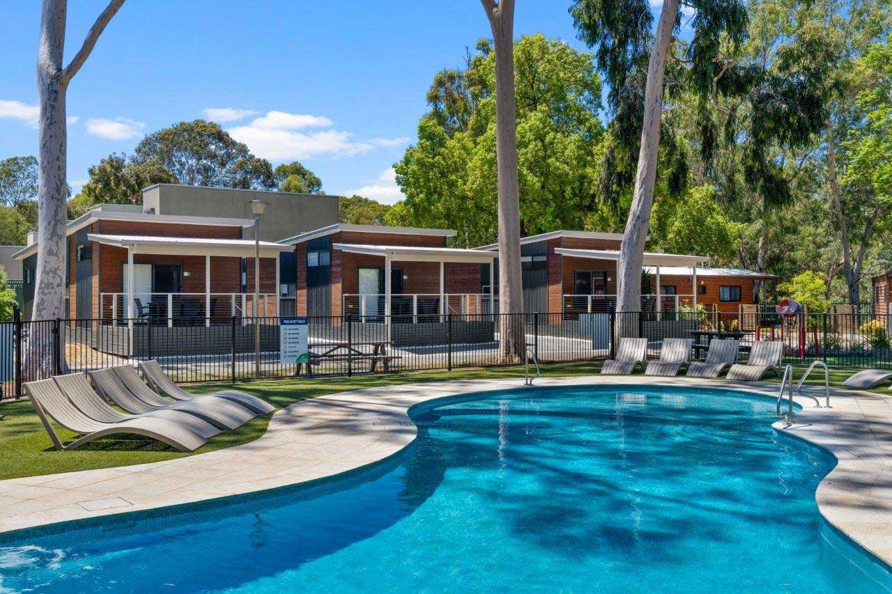 An outdoor swimming pool with lounge chairs around it, enclosed by a black fence, in front of modern apartment buildings surrounded by trees on a sunny day.