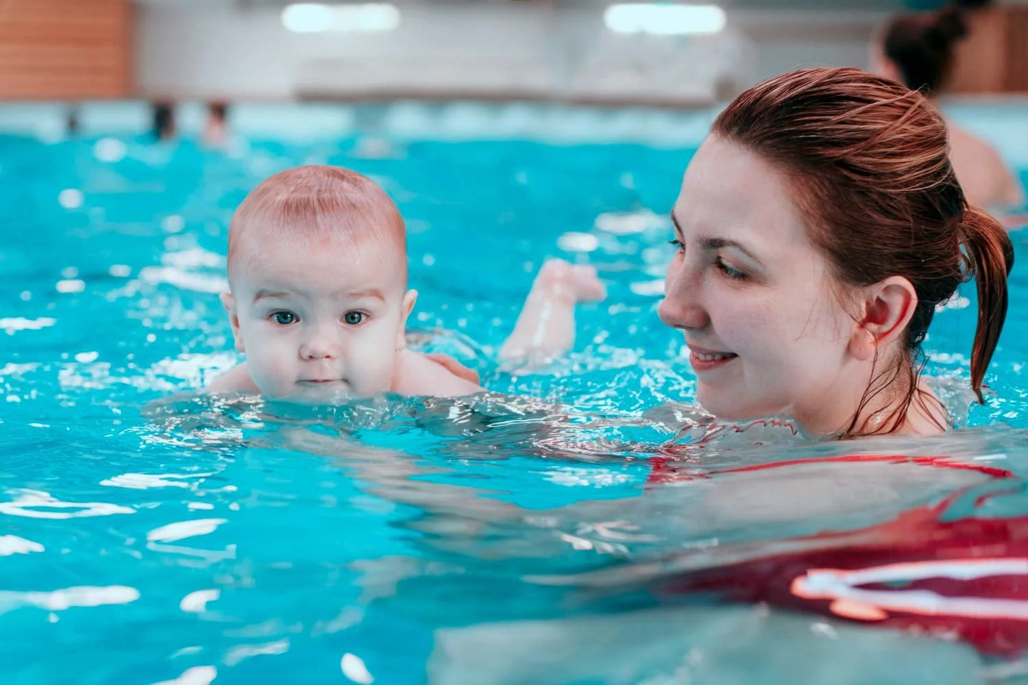 A woman is holding a baby in a swimming pool, with both of them partially submerged in water. The woman is smiling and the baby is looking directly at the camera.